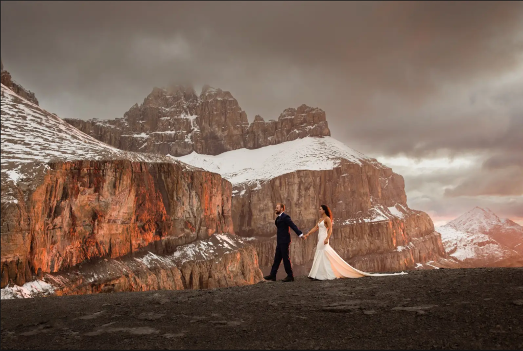 Bride and groom holding hands on a dramatic mountain peak in the Canadian Rockies at sunset — helicopter elopement with snow-capped cliffs, perfect for adventurous and unique destination weddings in Canada.