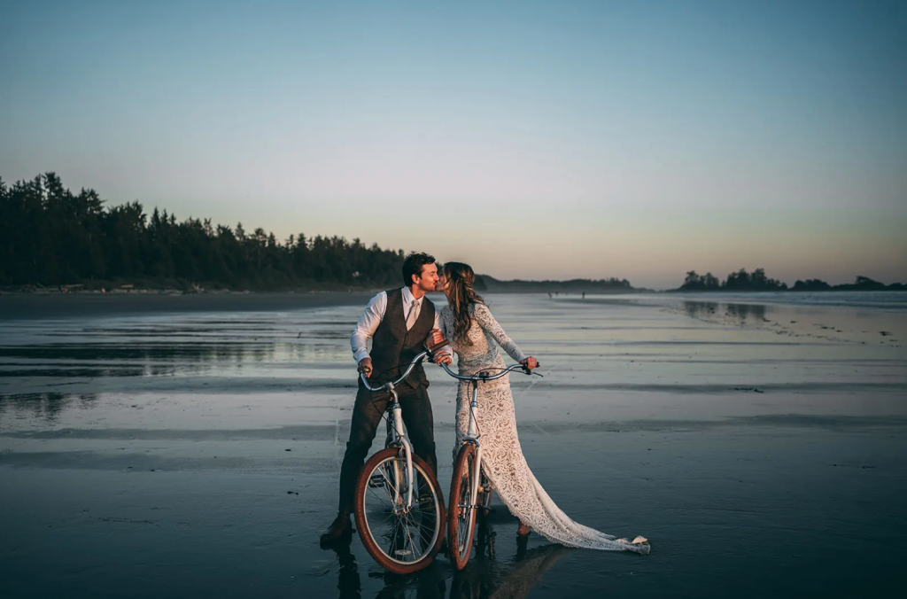 Bride and groom sharing a kiss on bicycles at sunset along Tofino’s sandy beach on Vancouver Island — relaxed coastal elopement capturing adventurous and unique destination wedding vibes in Canada.