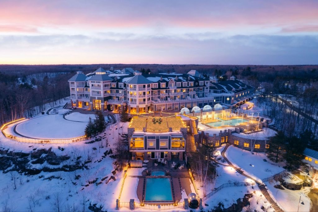 Aerial view of luxury lakeside resort in Muskoka, Ontario during winter sunset — glowing outdoor pools and snow-covered forest setting ideal for elegant and unique destination weddings in Canada.