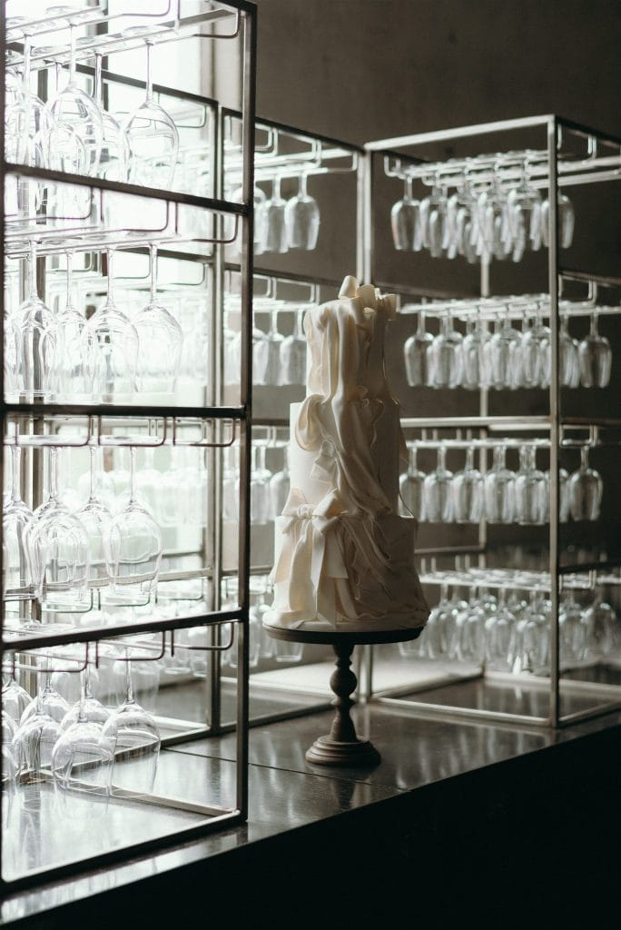 Elegant white multi-tiered wedding cake with delicate fabric-like fondant ruffles and bows, displayed on a wooden stand in a modern Toronto venue.