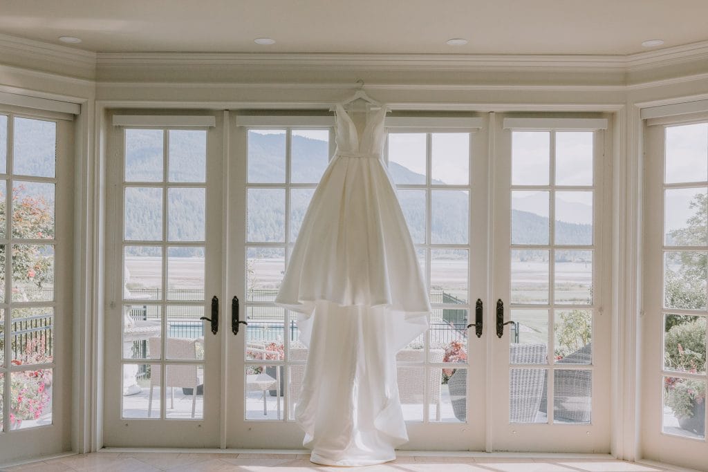 Classic A-line wedding dress hanging in front of large French doors with a mountain view backdrop, showcasing secondary lighting details and elegant bridal preparation.