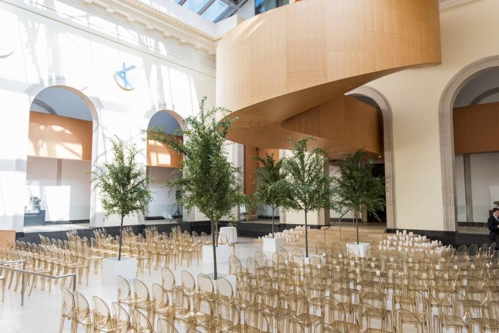 Modern wedding ceremony setup at the Art Gallery of Ontario (AGO) featuring ghost chairs, indoor trees, and the iconic sculptural wood spiral staircase in Toronto.