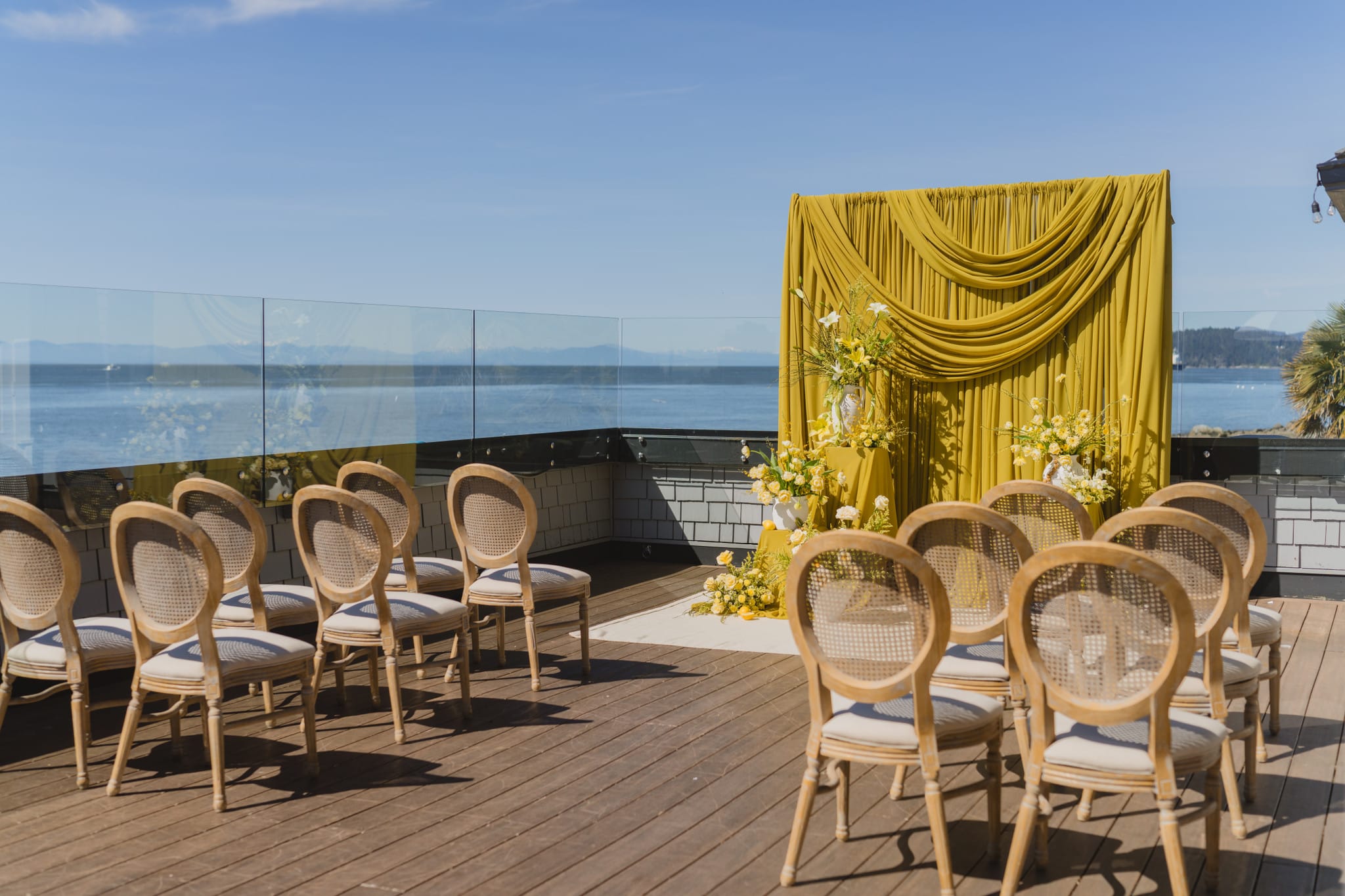Outdoor wedding ceremony setup on a terrace overlooking the ocean featuring a mustard yellow draped backdrop and elegant wooden chairs.
