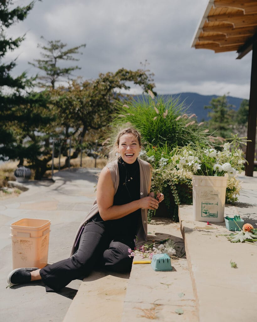 Smiling professional wedding florist arranging fresh white bouquets and greenery outdoors - Thistle & Thorne