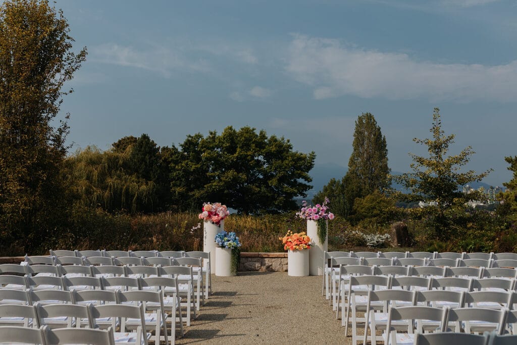 Scenic outdoor wedding ceremony venue with rows of white chairs and colourful floral pillars - Thistle & Thorne
