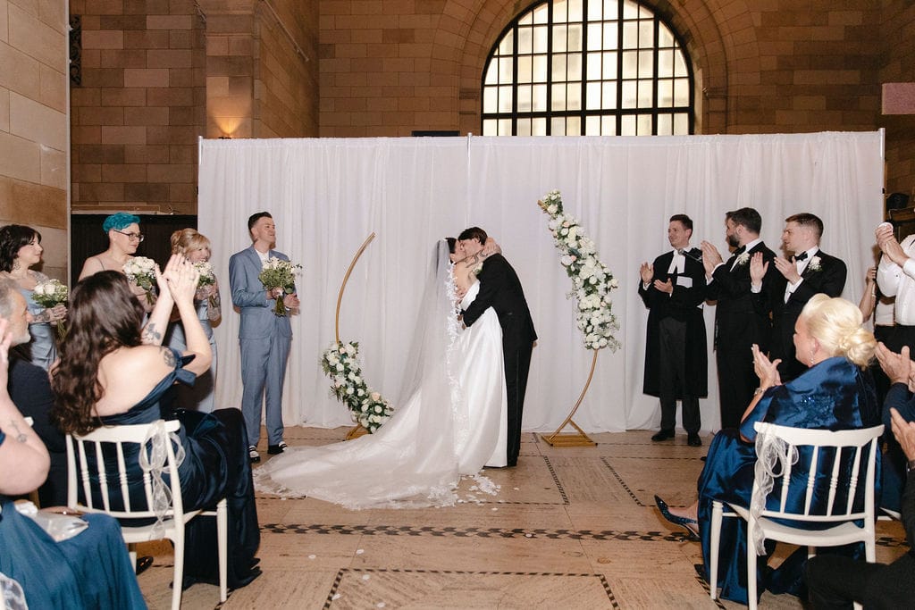 A bride and groom share their first kiss under a minimalist gold floral arch during an indoor wedding ceremony at 1868 building, illustrating boutique Montreal wedding cost elegance - Thistle & Thorne