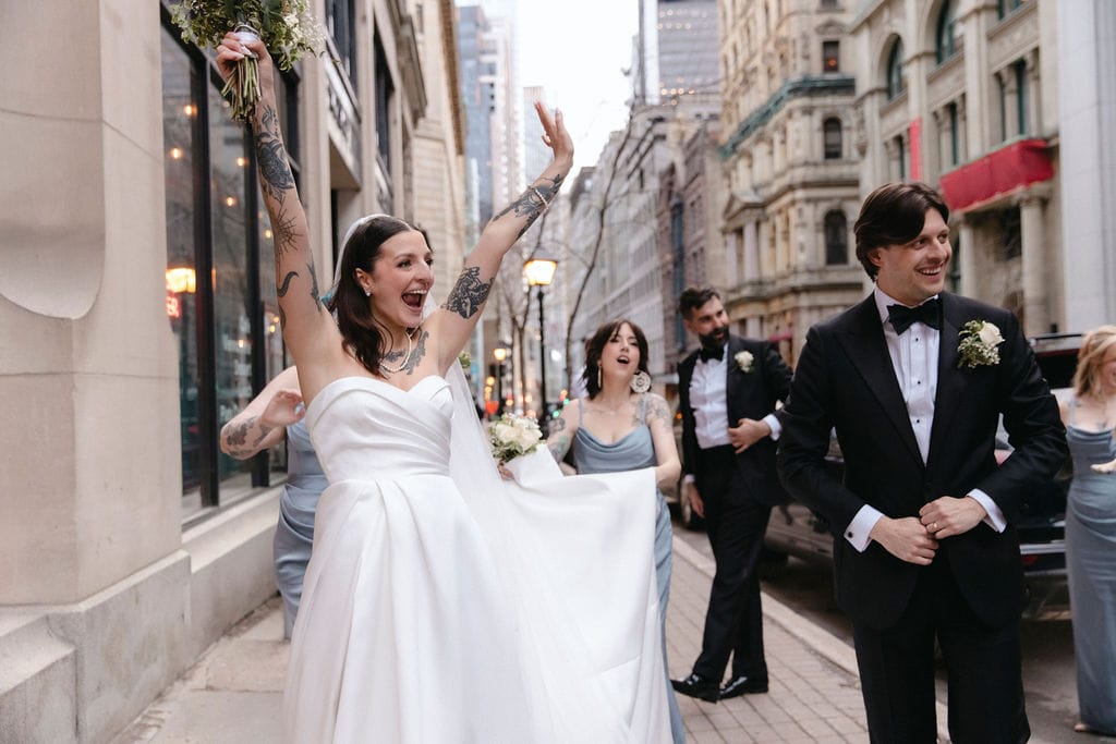 Exuberant newly married couple celebrating on a city street, bride holding bouquet high, showcasing urban wedding style and joy, illustrative of wedding cost planning by Thistle & Thorne.