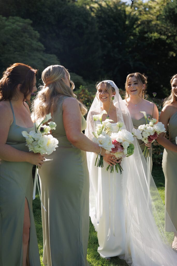 Smiling bride with bridesmaids in sage green dresses holding white flower bouquets at an outdoor ceremony - Thistle & Thorne