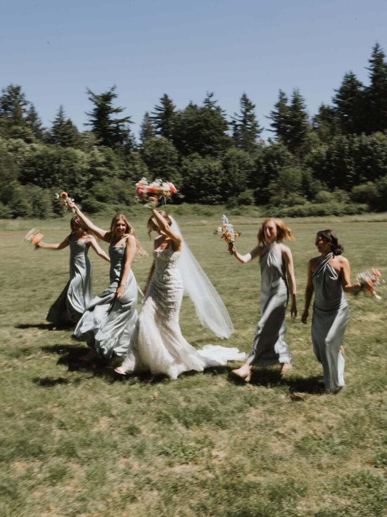 Joyful bride and bridesmaids running through a grassy field while holding their bouquets - Thistle & Thorne